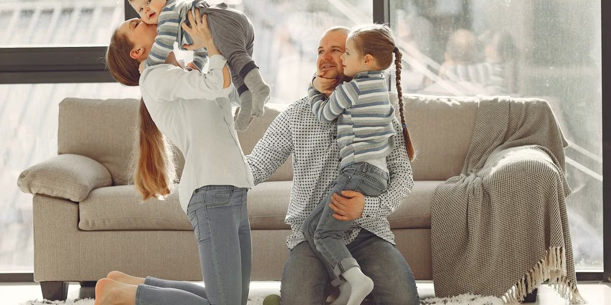 Cheerful mother kissing little boy while father hugging adorable daughter when family sitting on floor in rainy day against window