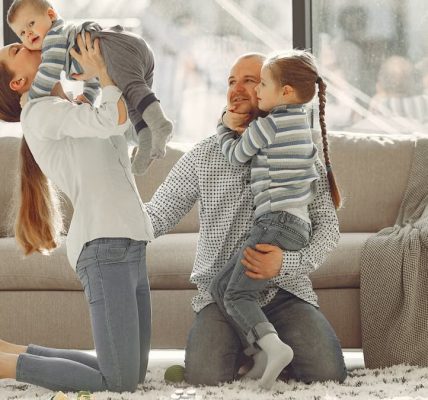 Cheerful mother kissing little boy while father hugging adorable daughter when family sitting on floor in rainy day against window