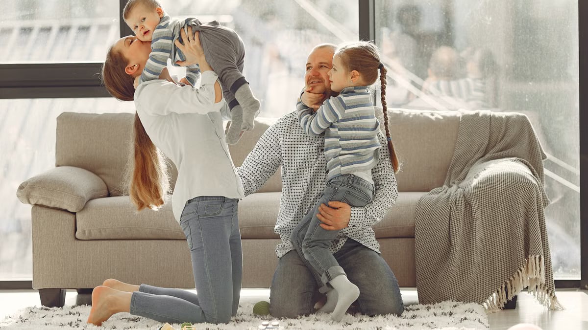 Cheerful mother kissing little boy while father hugging adorable daughter when family sitting on floor in rainy day against window